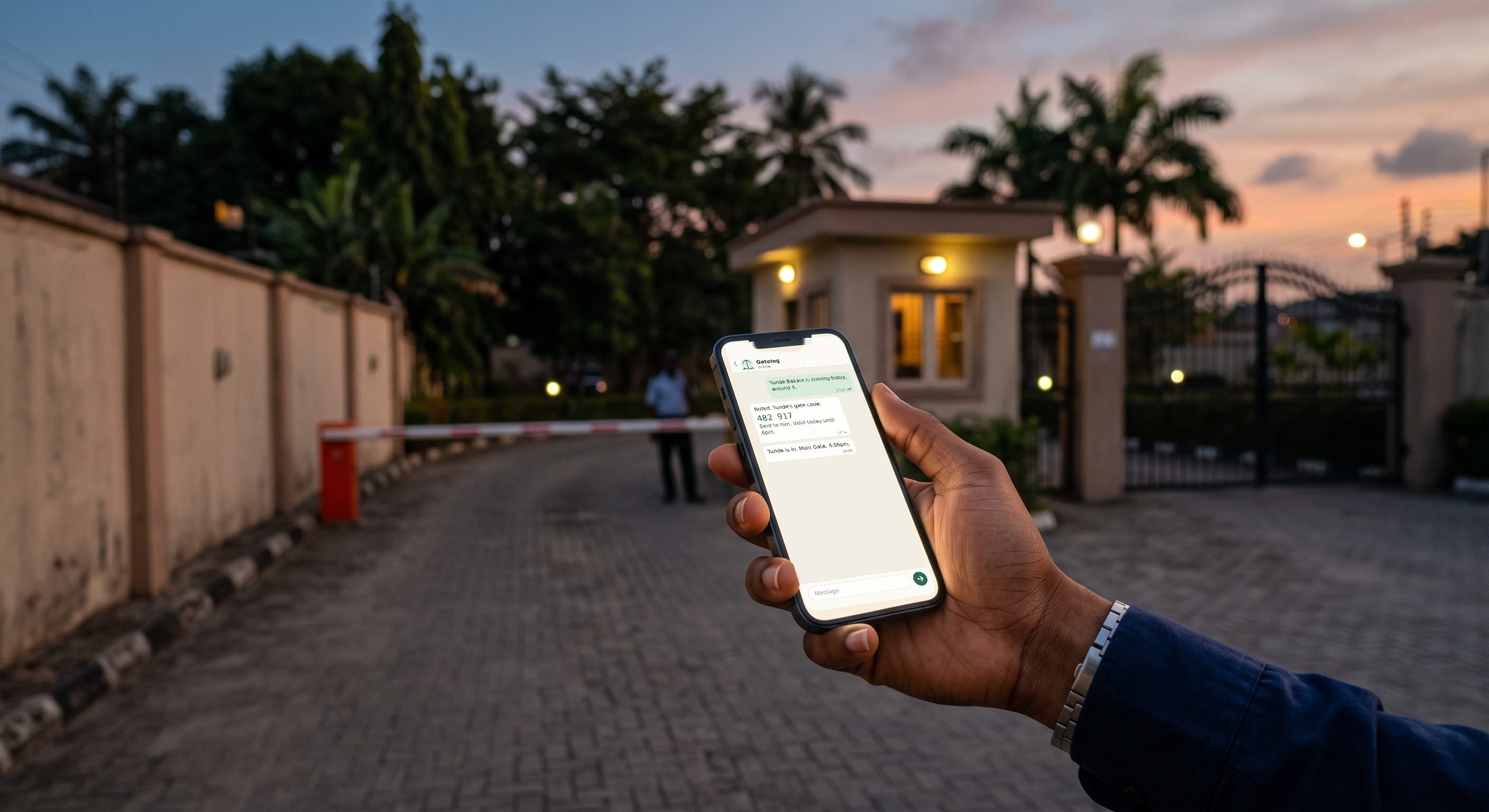 A resident holds a phone at the entrance of a gated estate at dusk. On screen, the Gatelog chat: a guest announced, a gate code issued, and the arrival confirmed.
